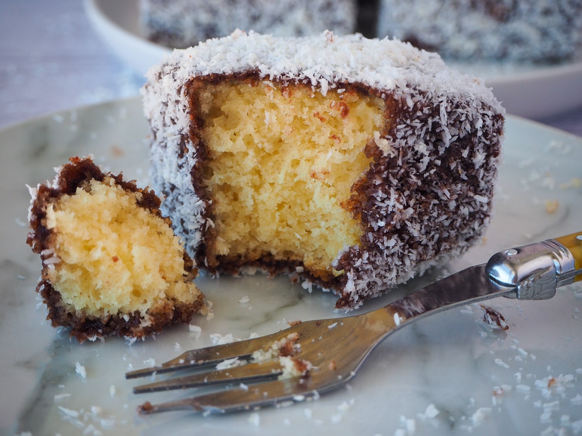 Close up of lamington with a piece taken out with a fork and vintage cake for next to it.
