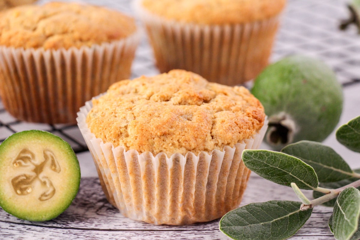 Feijoa muffins with fresh feijoas and feijoa leaves, with extra muffins on a cooling rack in the background.