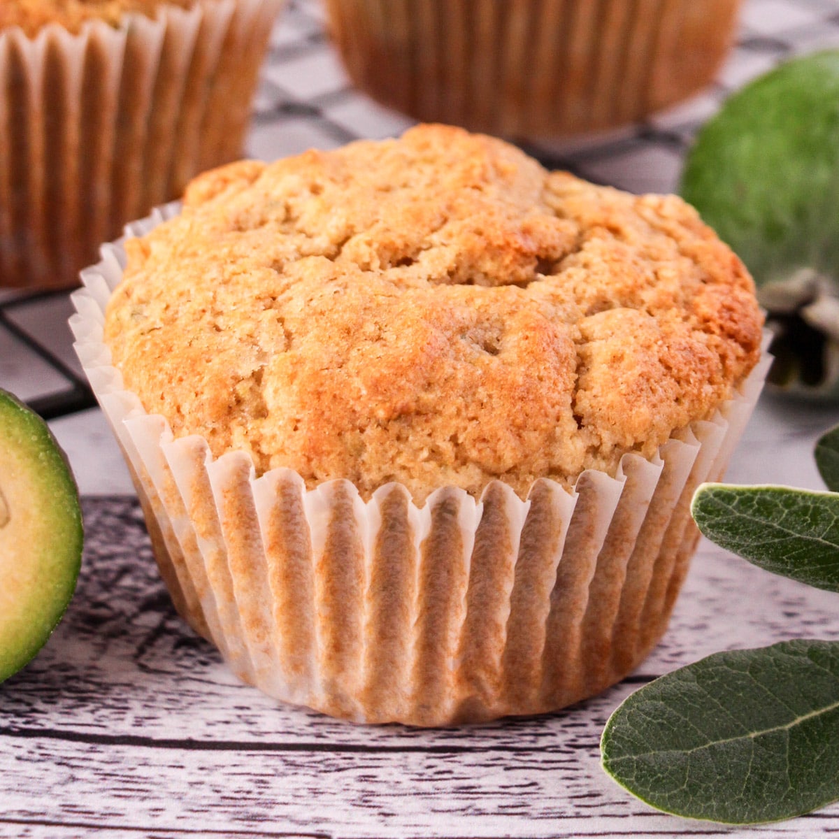 Close up feijoa muffins with fresh feijoas and feijoa leaves, with extra muffins on a cooling rack in the background.