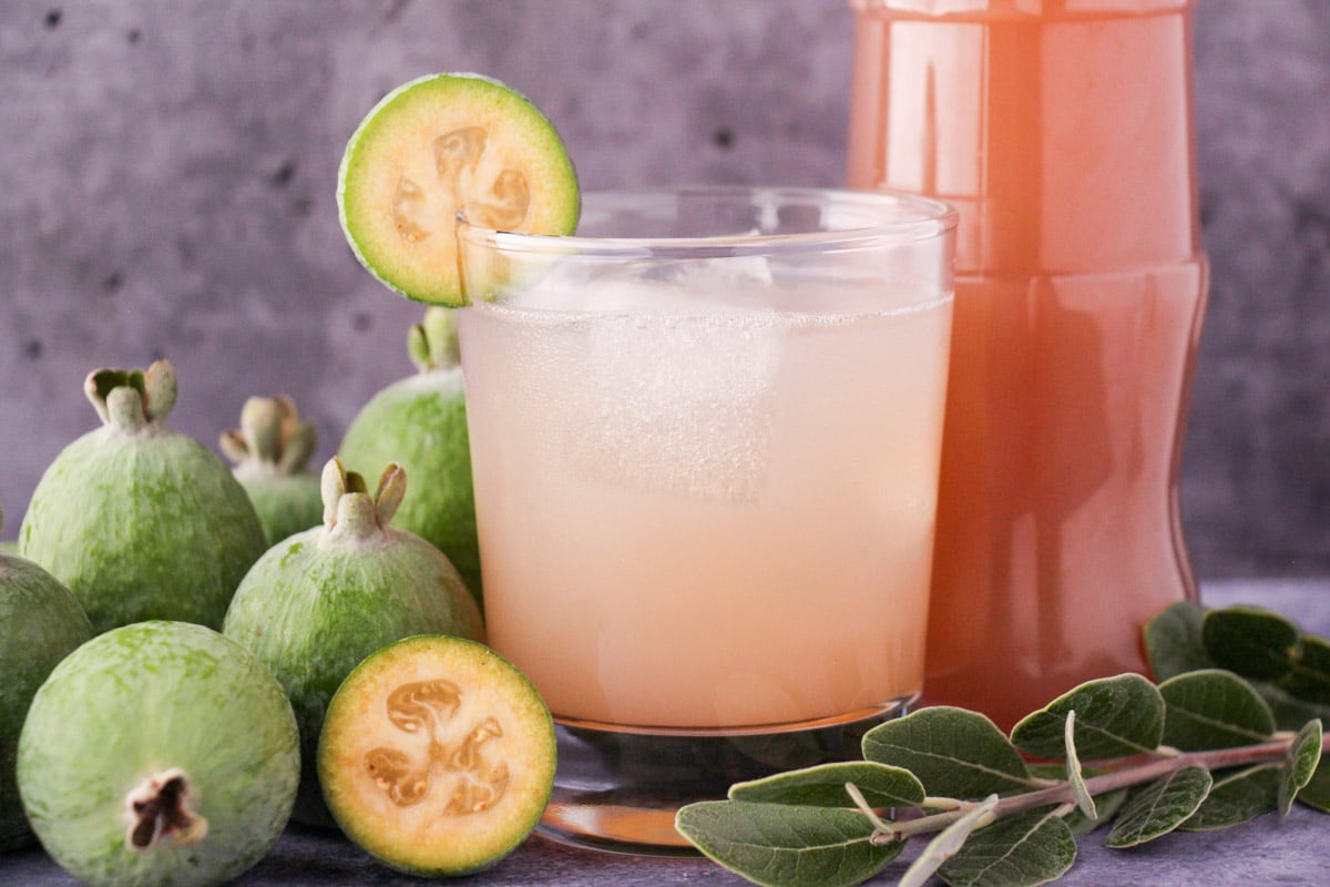 Feijoa syrup 10 Glass of made up feijoa syrup with ice and fresh feijoa garnish, with bottle of feijoa syrup and fresh feijoas and leaves.