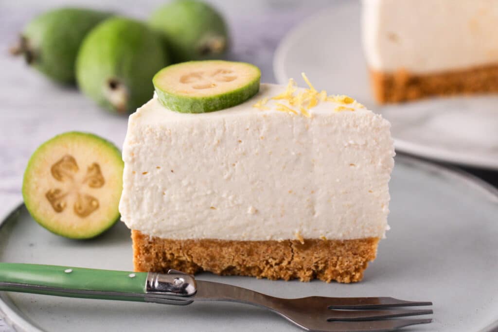 Feijoa cheesecake with slice taking out, silver serving ware and fresh feijoas and feijoa leaves in the background.