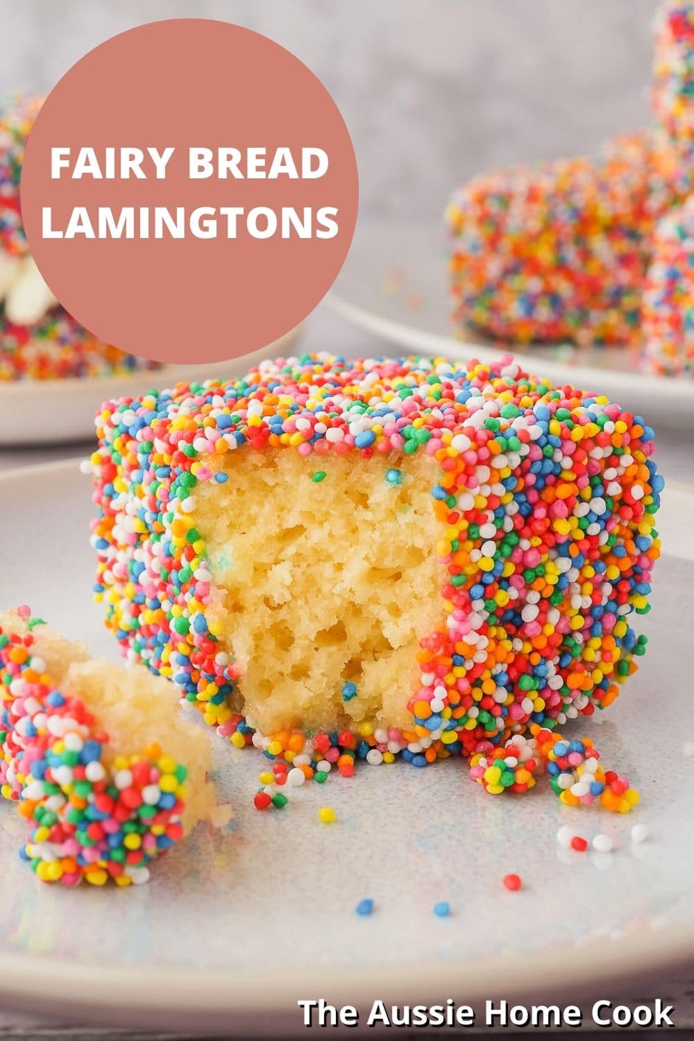 Lamington on a plate with one piece out, with lamingtons in the background and text overlay, Fairy Bread Lamingtons and The Aussie Home Cook.