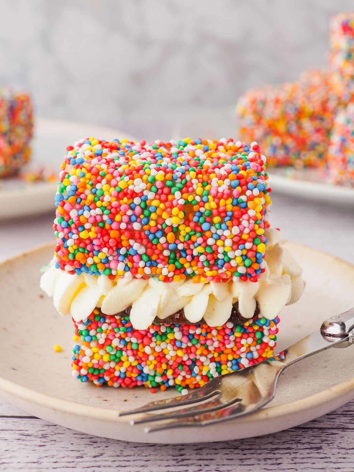 Lamington with jam and cream on a plate with a fork, and plate of lamingtons in the backgroun.