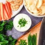 Sour cream dip garnished with fresh chives, with a bowl of chips, plate of crackers and carrot sticks, and chopping board with freshly chopped chives.